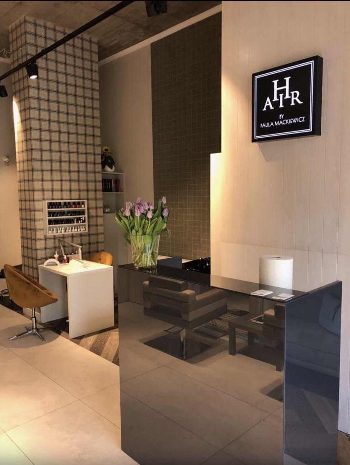 Nowoczesne wnętrze salonu, z recepcją o lustrzanym wykończeniu, stanowiskiem do manicure z paletą lakierów, krzesłem w odcieniu musztardowym oraz logo salonu na ścianie.
