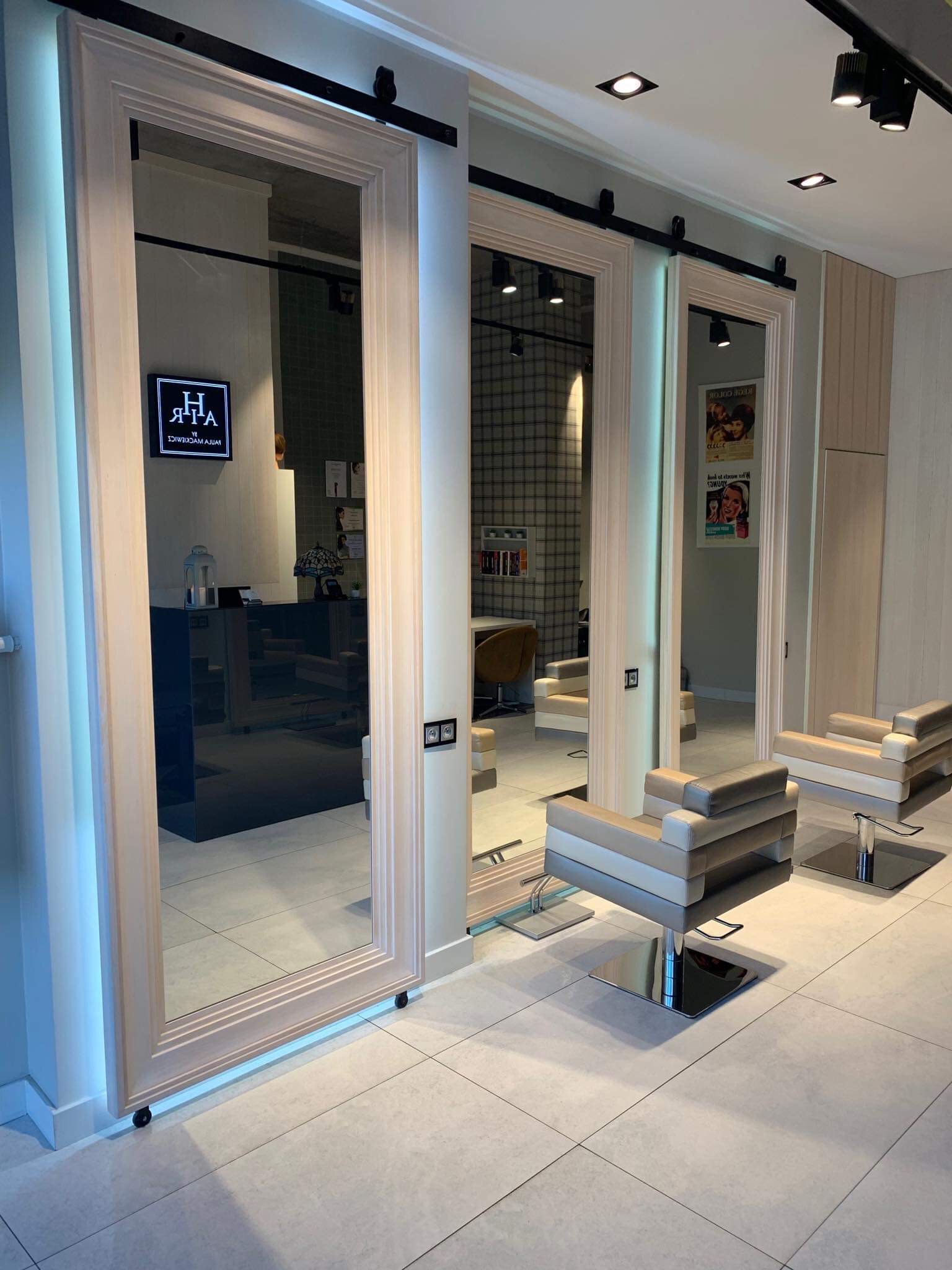 Wnętrze salonu z trzema lustrami na szynach, fotelami fryzjerskimi w odcieniach beżu i szarości oraz jasną podłogą z płytek.