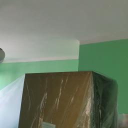 M&M Firma Usługowa Gładzie i Malowanie - Pokój w trakcie malowania na zielono, meble zabezpieczone folią, widoczny żyrandol i okno z otwartymi skrzydłami.