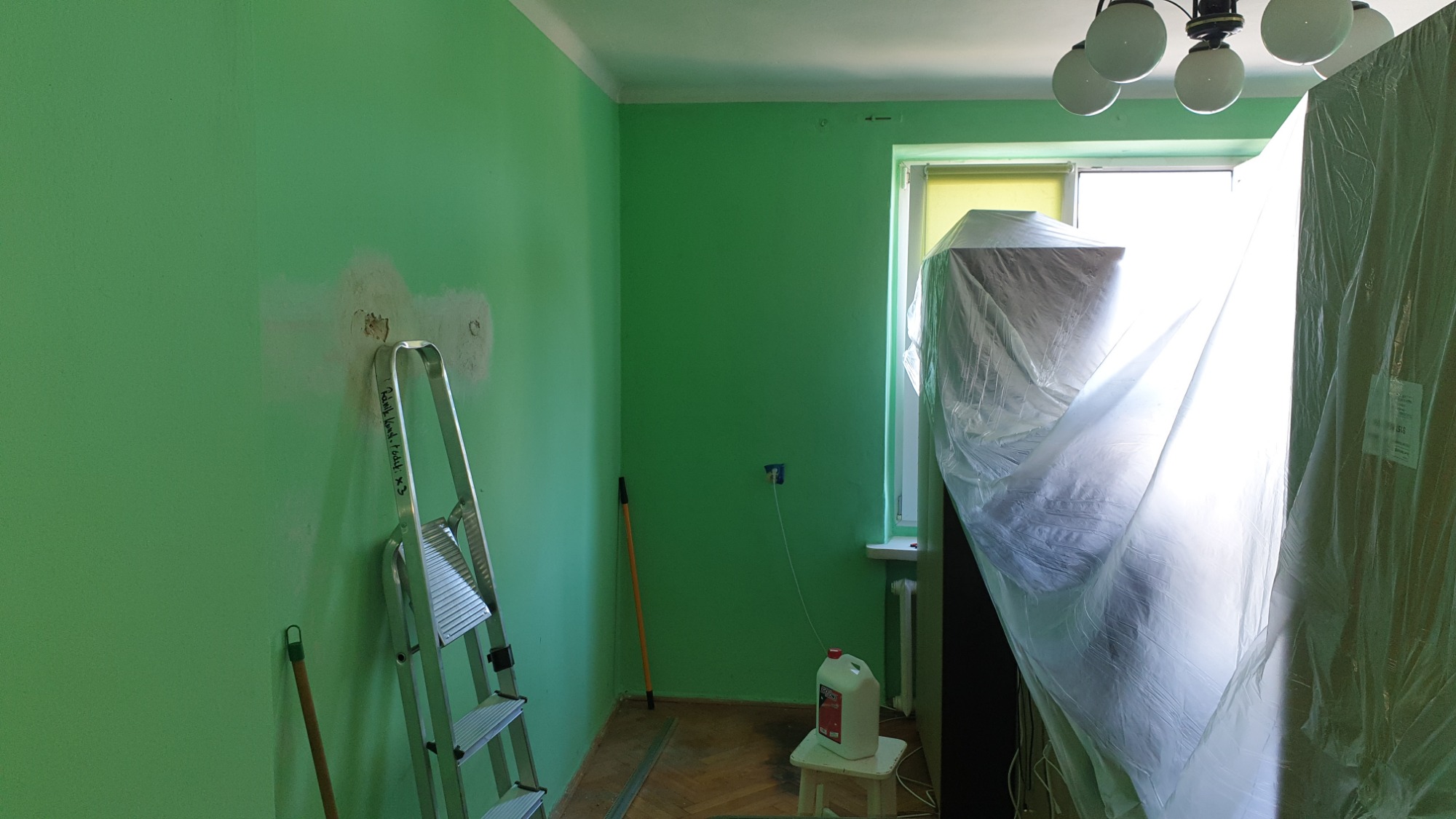 Pokój w trakcie odświeżania z zielonymi ścianami, zabezpieczone meble folią, drabina oparta o ścianę z widocznymi śladami po naprawie ubytków, podłoga parkietowa, butelka z preparatem na taborecie.