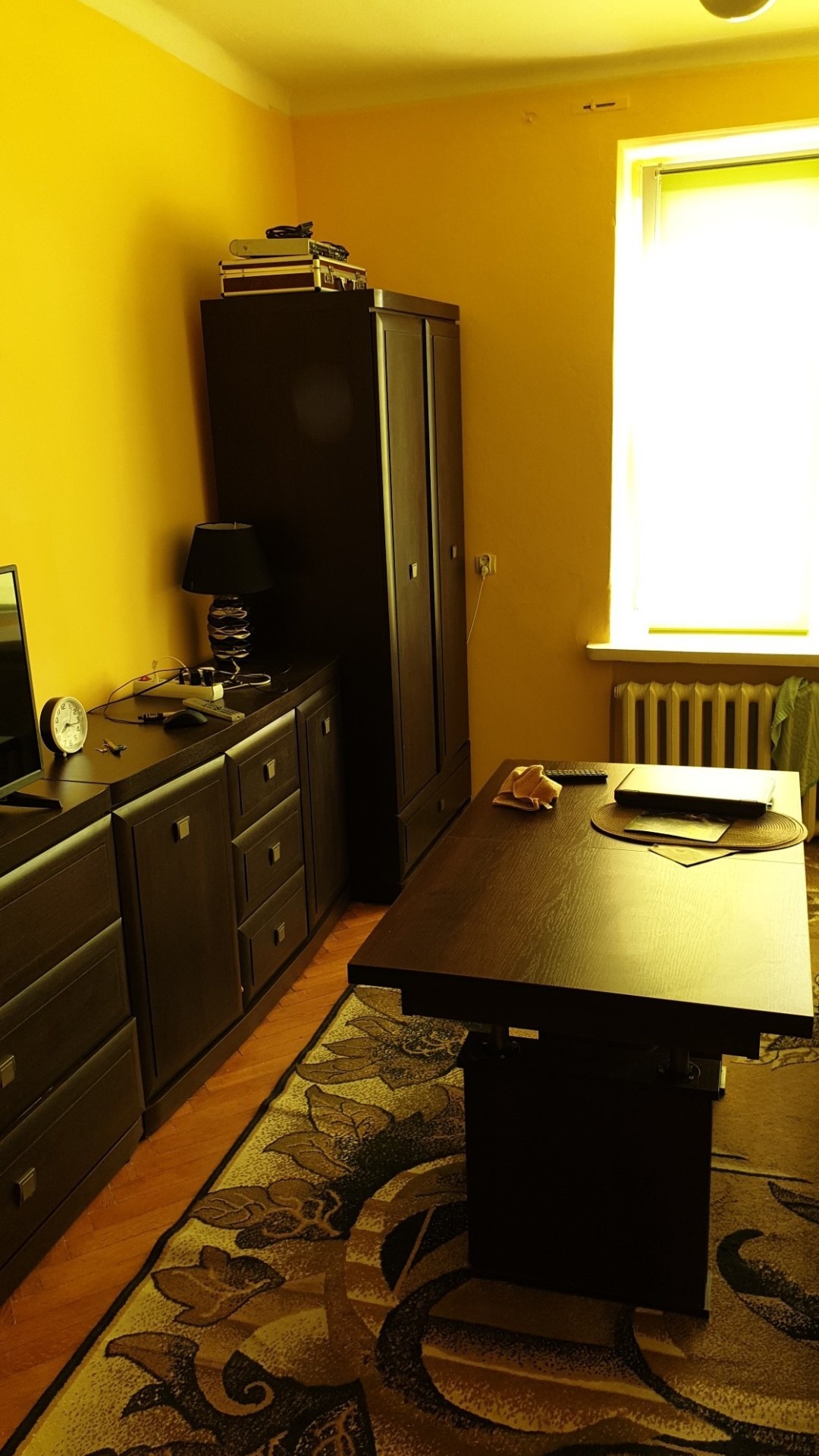 Pokój z żółtymi ścianami, ciemnymi meblami, rozłożonym stołem z laptopem i wzorzystym dywanem.