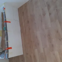 Układanie paneli podłogowych w trakcie remontu, widoczne zabezpieczenia ścian i narożników taśmą malarską oraz wiadro z zaprawą.