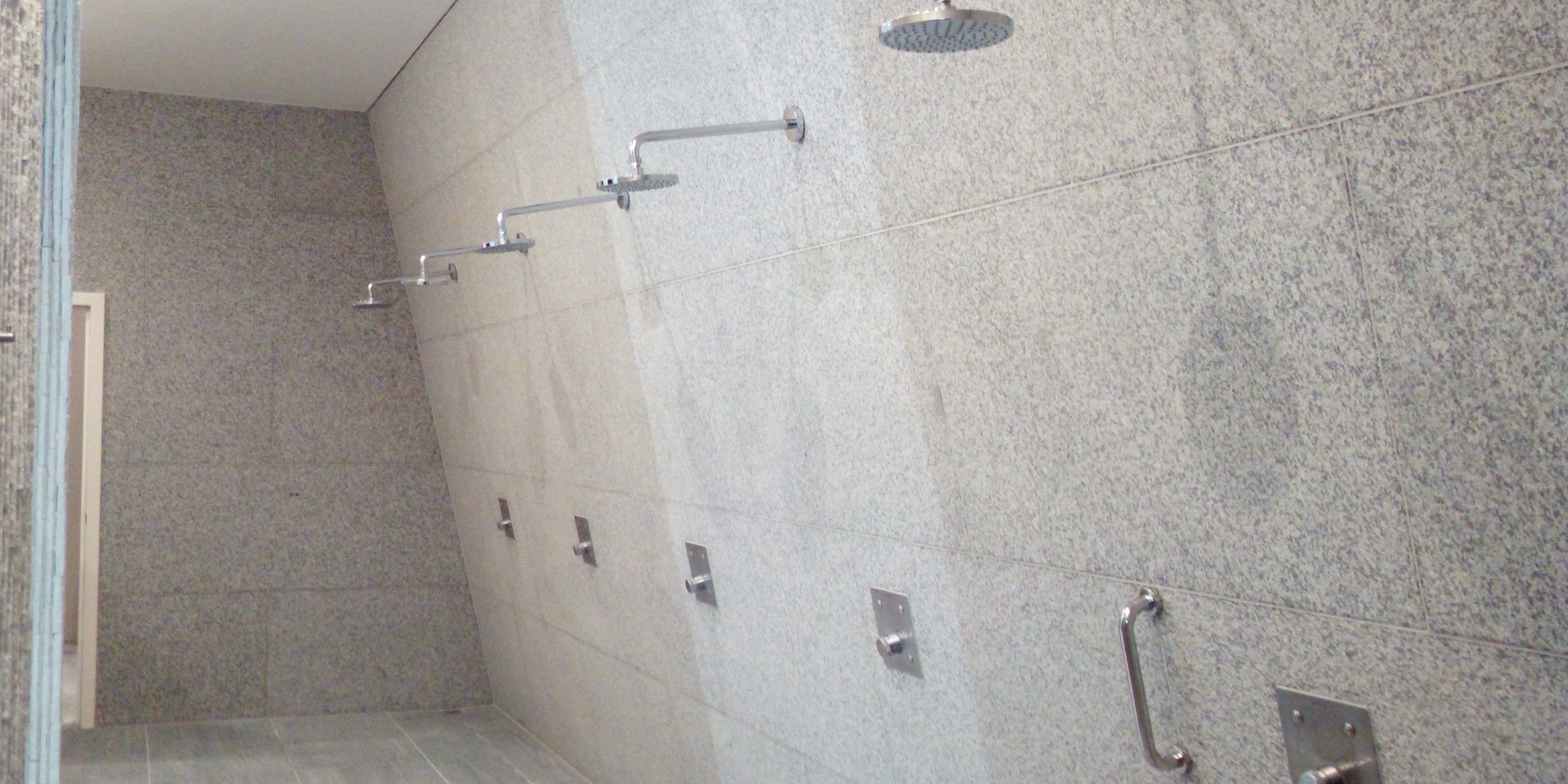 Publiczna łazienka z kabinami prysznicowymi wyłożonymi szarymi płytkami imitującymi kamień, widoczne chromowane słuchawki prysznicowe i uchwyty dla osób z niepełnosprawnościami.