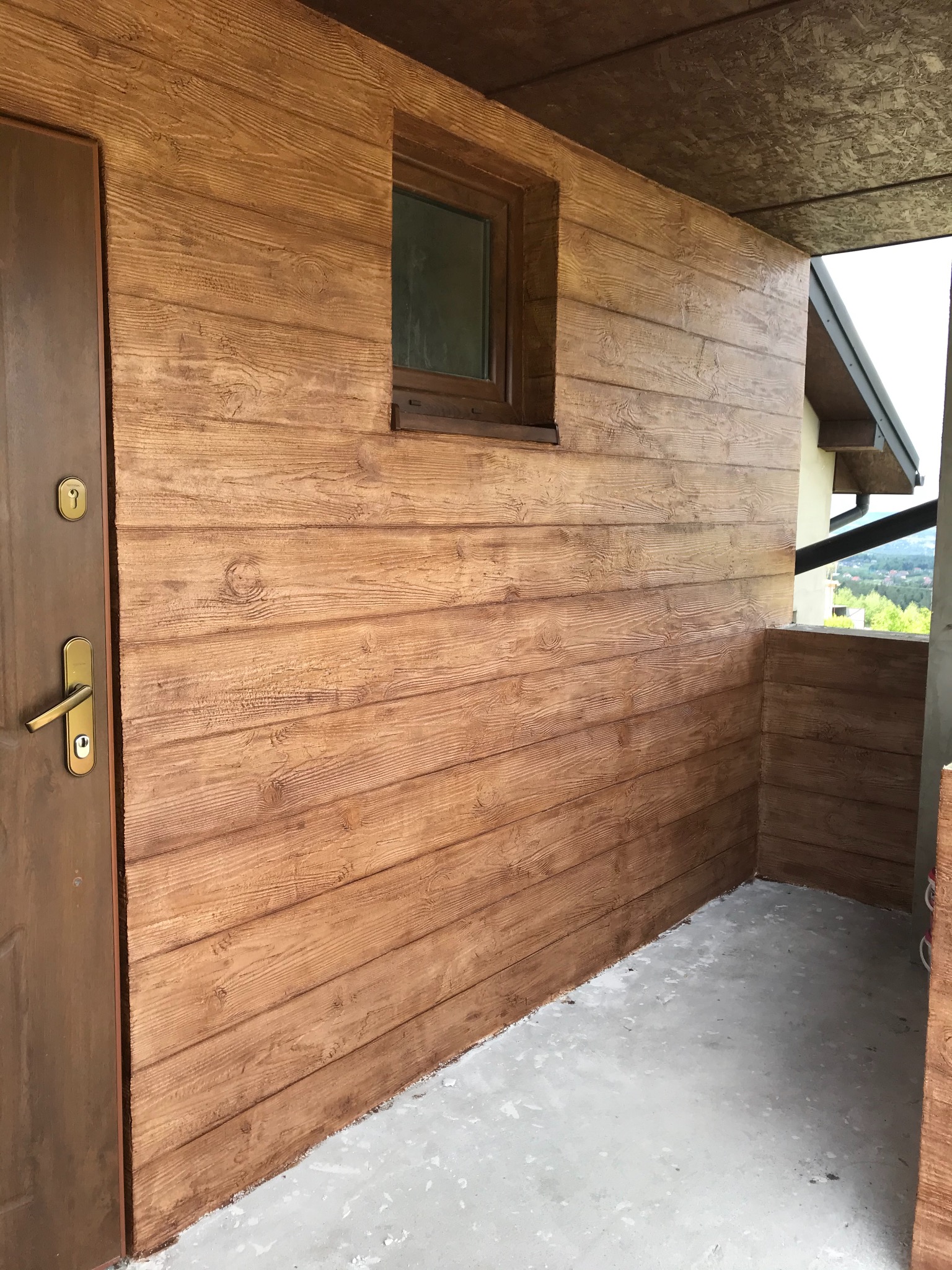 Brązowa elewacja budynku imitująca drewno, z widocznymi słojami, małe okno i drzwi wejściowe z mosiężną klamką, betonowa podłoga werandy.