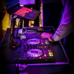 DJ pracujący na profesjonalnym sprzęcie: mikser, laptop z podświetlaną klawiaturą, mikrofon. Scena oświetlona fioletowym światłem, widoczny lanyard z napisem 'DJ Bakcyl'.