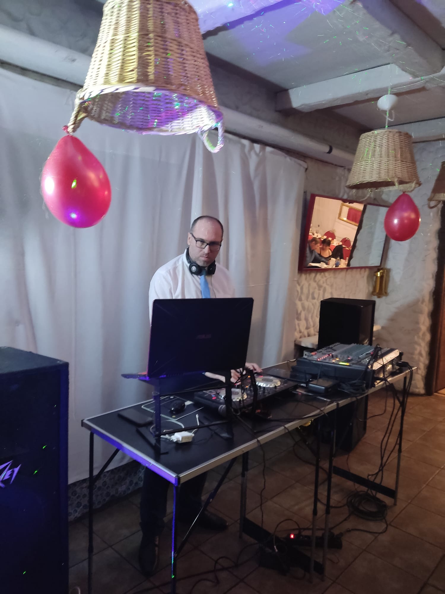 DJ z laptopem i mikserem na tle dekoracji z wiklinowych koszy i czerwonych balonów w pomieszczeniu z widocznym odbiciem imprezy w lustrze.