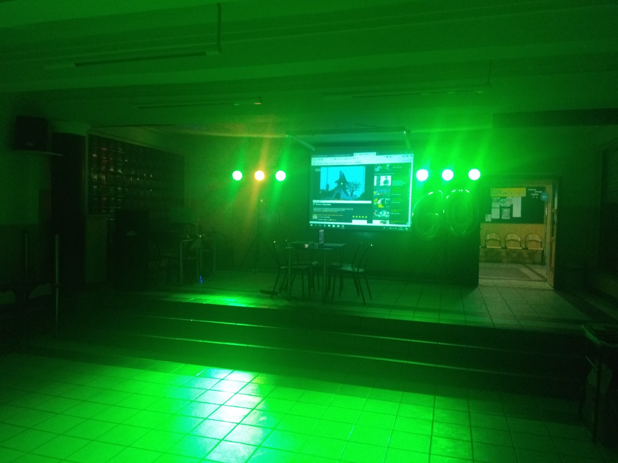 Scena oświetlona zielonym światłem z projektorem wyświetlającym ekran komputera, na scenie stolik z krzesłami.