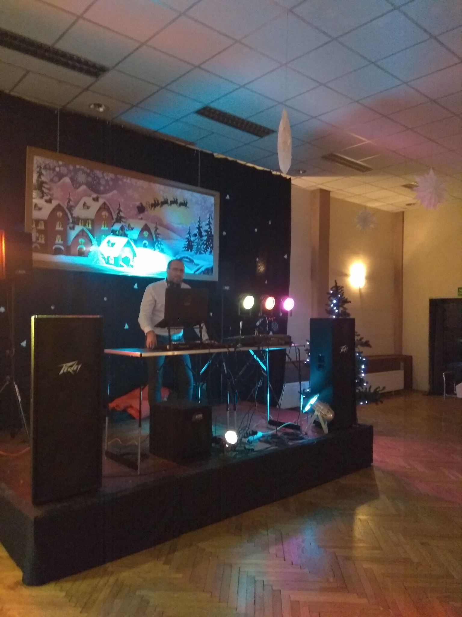 DJ z laptopem i sprzętem nagłośnieniowym na podwyższeniu, w tle świąteczna dekoracja z motywem sań Świętego Mikołaja i oświetlenie sceniczne.