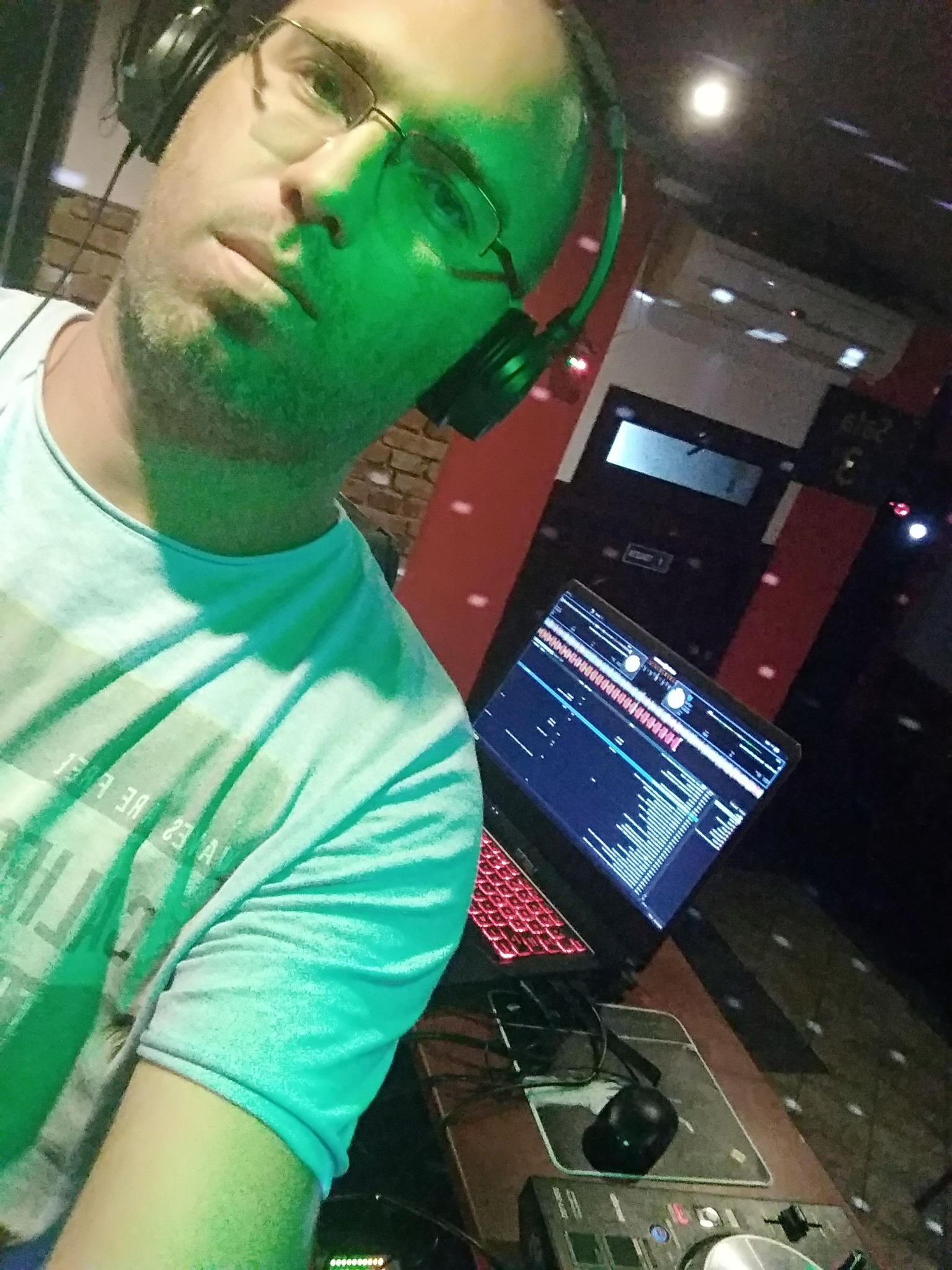 Mężczyzna w okularach i słuchawkach, oświetlony zielonym światłem, stoi za konsoletą DJ-ską z laptopem wyświetlającym oprogramowanie muzyczne, w tle widoczne elementy dekoracyjne sali.