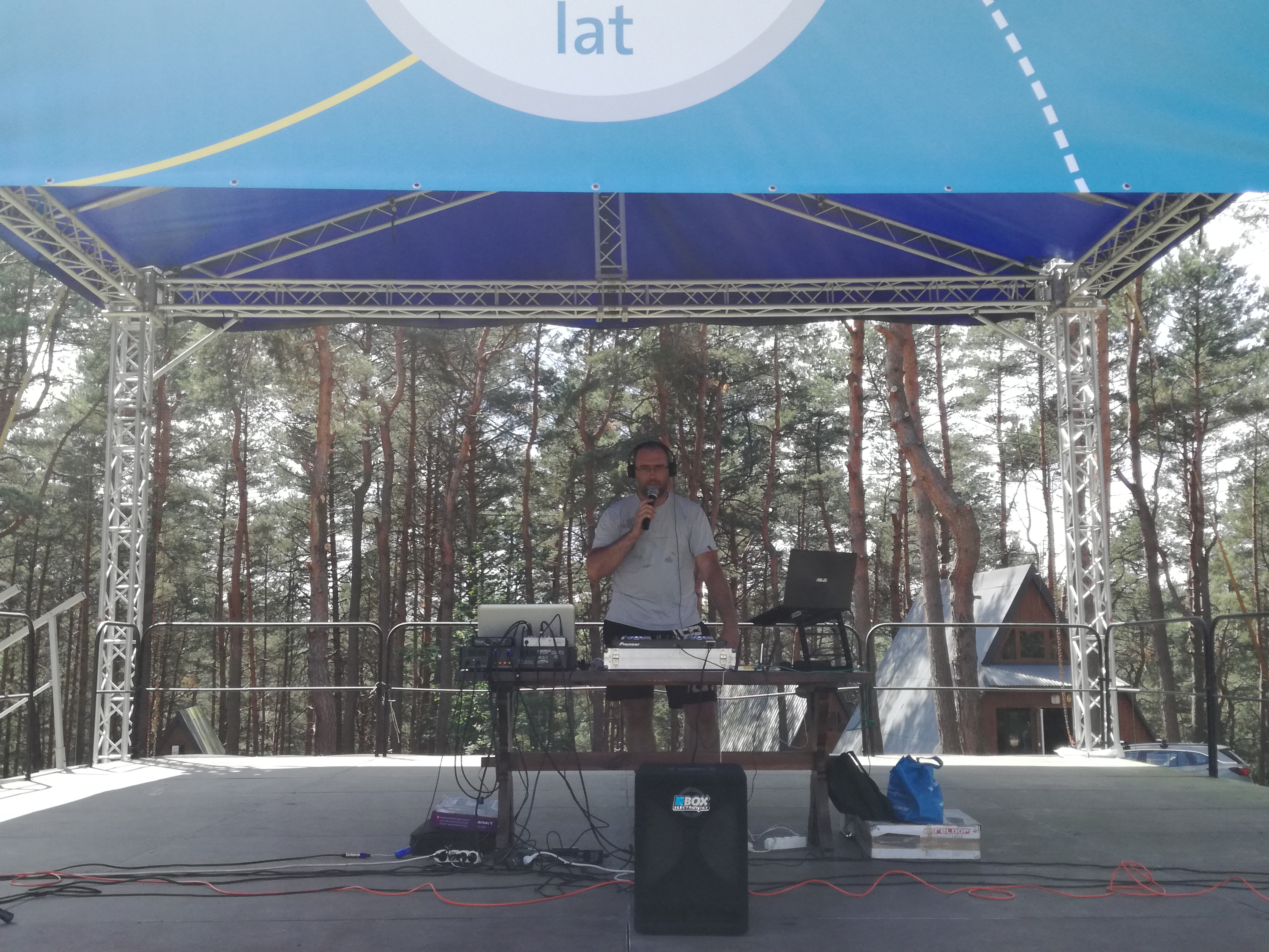 DJ w słuchawkach i z mikrofonem na scenie plenerowej z nagłośnieniem, laptopem i lasem w tle.