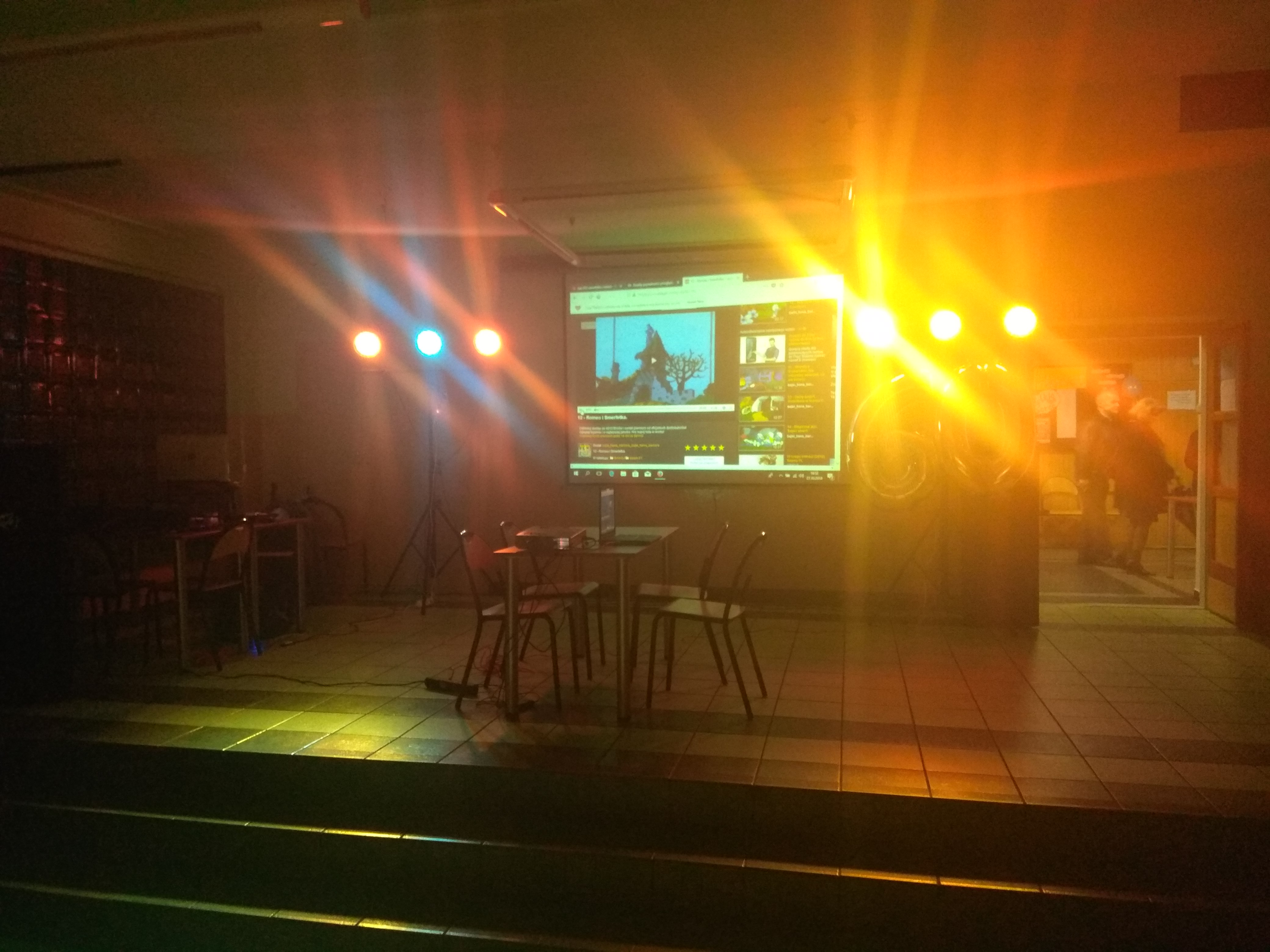 Sala z oświetleniem scenicznym, projektorem wyświetlającym ekran komputera, stolikami i krzesłami. Widoczne osoby w tle.