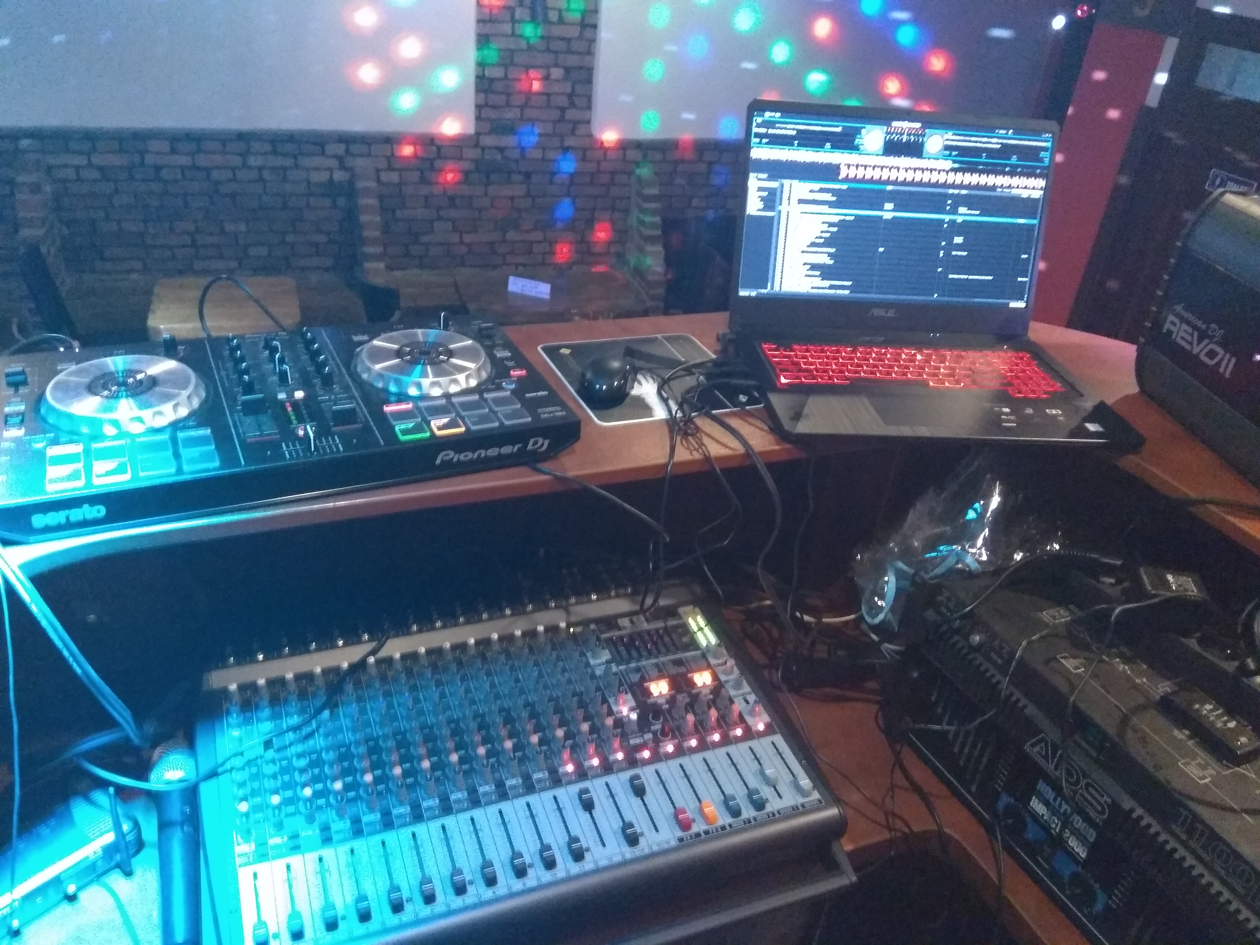 DJ stanowisko z mikserem, kontrolerem Pioneer DJ, laptopem z czerwonym podświetleniem klawiatury i oświetleniem dyskotekowym w tle.