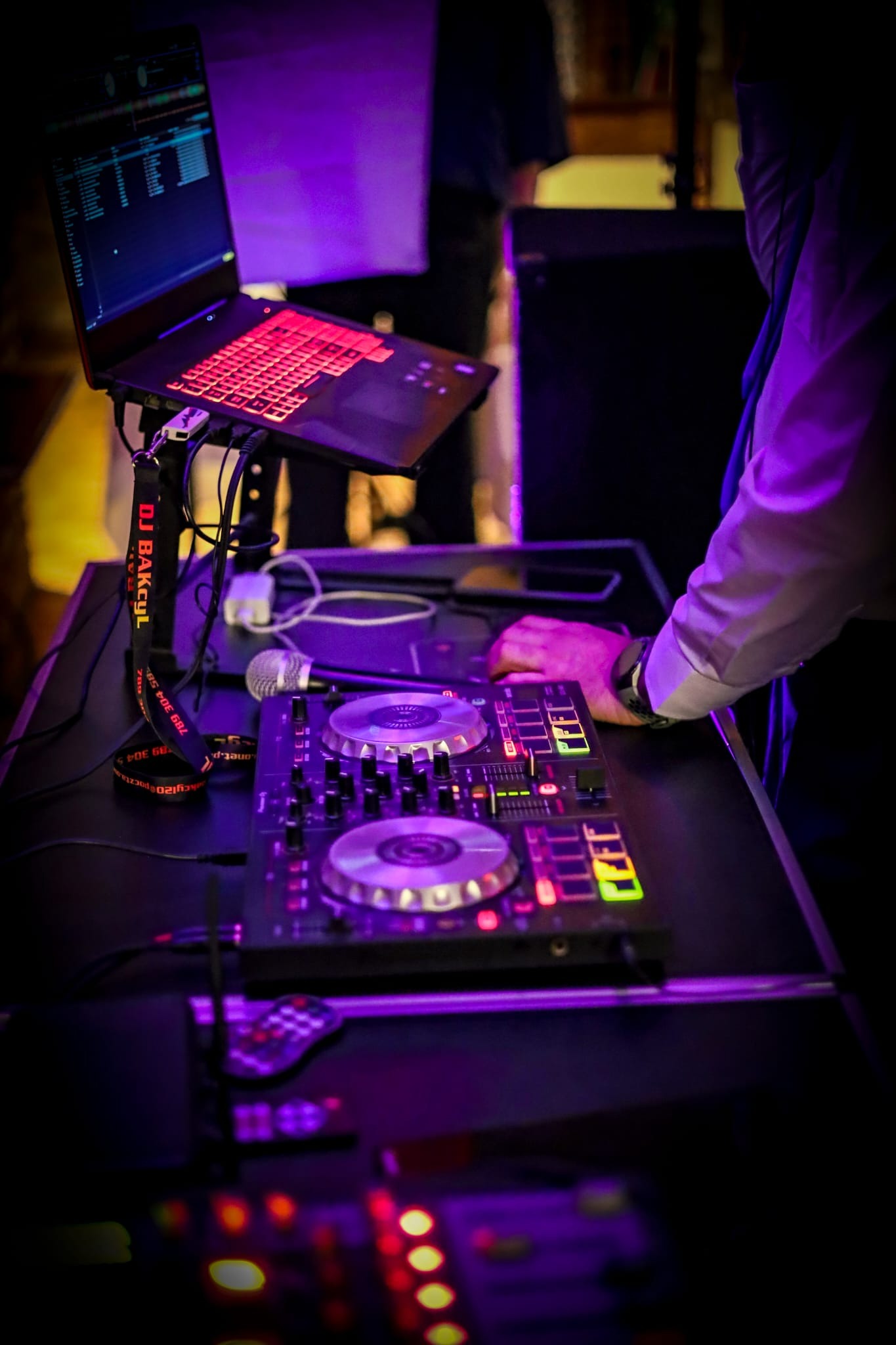 DJ pracujący na profesjonalnym sprzęcie: mikser, laptop z podświetlaną klawiaturą, mikrofon. Scena oświetlona fioletowym światłem, widoczny lanyard z napisem 'DJ Bakcyl'.