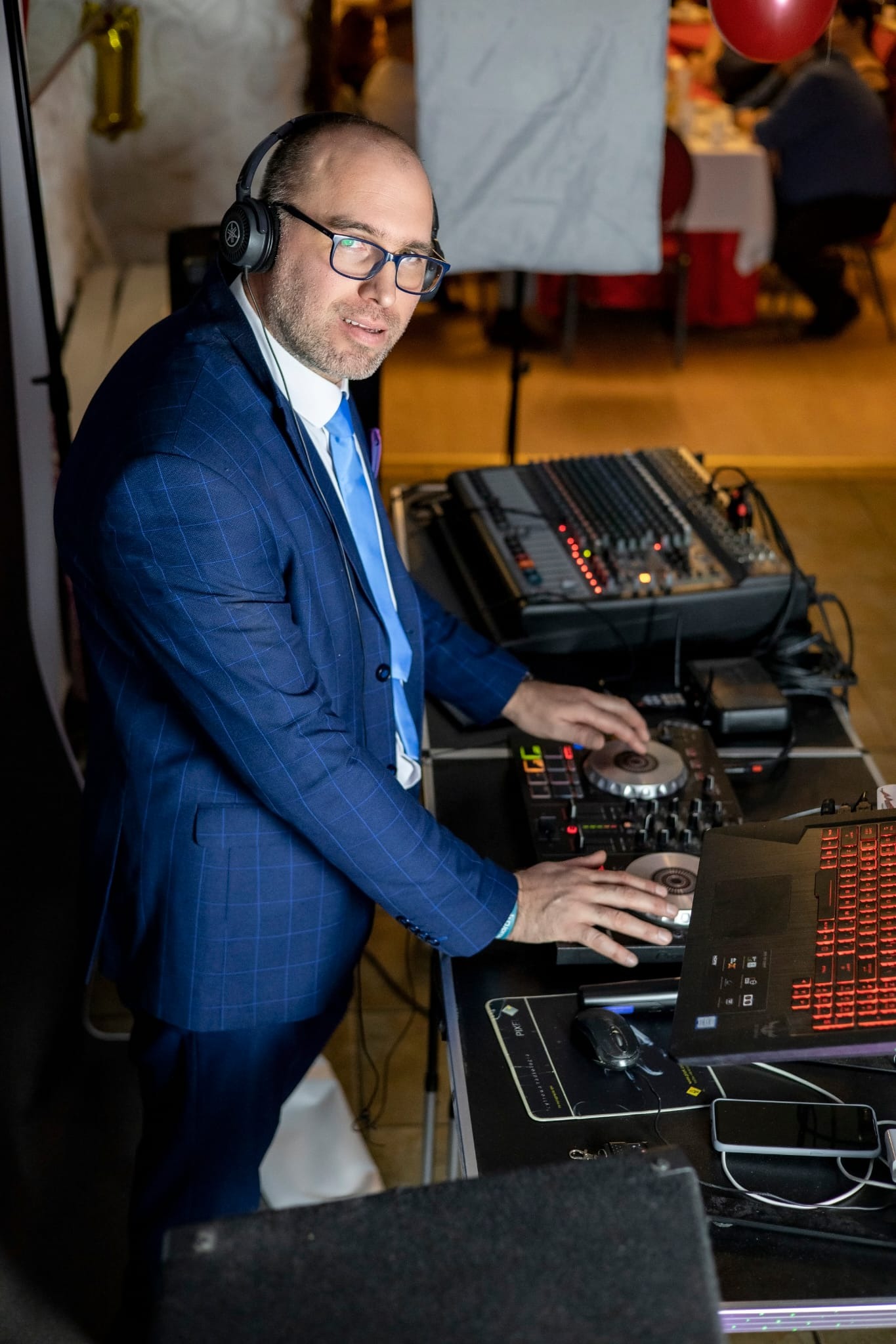 DJ w niebieskim garniturze i słuchawkach, miksujący muzykę na konsoli podczas imprezy z czerwonymi balonami w tle.
