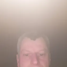 Portret mężczyzny z bliska, z wyraźnymi rysami twarzy i krótkimi, siwiejącymi włosami, na rozmytym, ciemnym tle. Oświetlenie od góry.