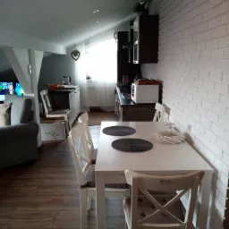 Jasne wnętrze na poddaszu z białym stołem i krzesłami, dekoracyjną ścianą z białej cegły, oraz widokiem na aneks kuchenny i salon z telewizorem.