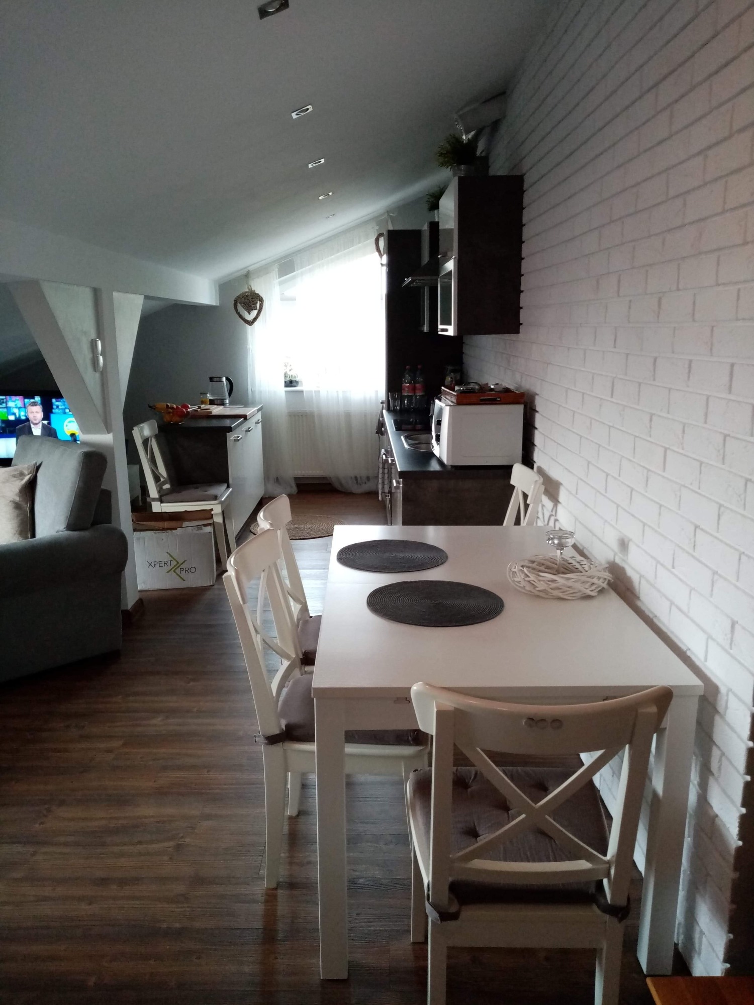 Jasne wnętrze na poddaszu z białym stołem i krzesłami, dekoracyjną ścianą z białej cegły, oraz widokiem na aneks kuchenny i salon z telewizorem.