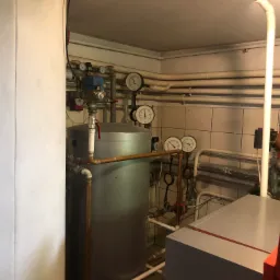 Wnętrze kotłowni z widocznym srebrnym zbiornikiem buforowym, licznymi rurami, manometrami i kotłem grzewczym Viessmann, całość na tle kwadratowych płytek ściennych.