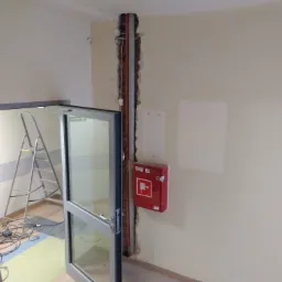 Wnęka w ścianie z widocznymi rurami instalacyjnymi, obok czerwona szafka hydrantowa, drabina i rozłożone kable na podłodze w tle, widok przez otwarte szklane drzwi.