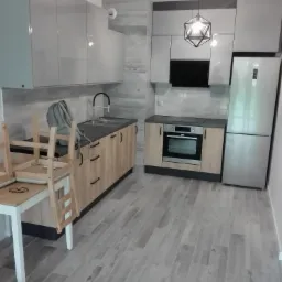 Jasna kuchnia z drewnianymi frontami szafek dolnych i szarymi górnymi, z blatem w kolorze antracytowym, oświetlona geometryczną lampą wiszącą, z krzesłem i okularami na stole w rogu.