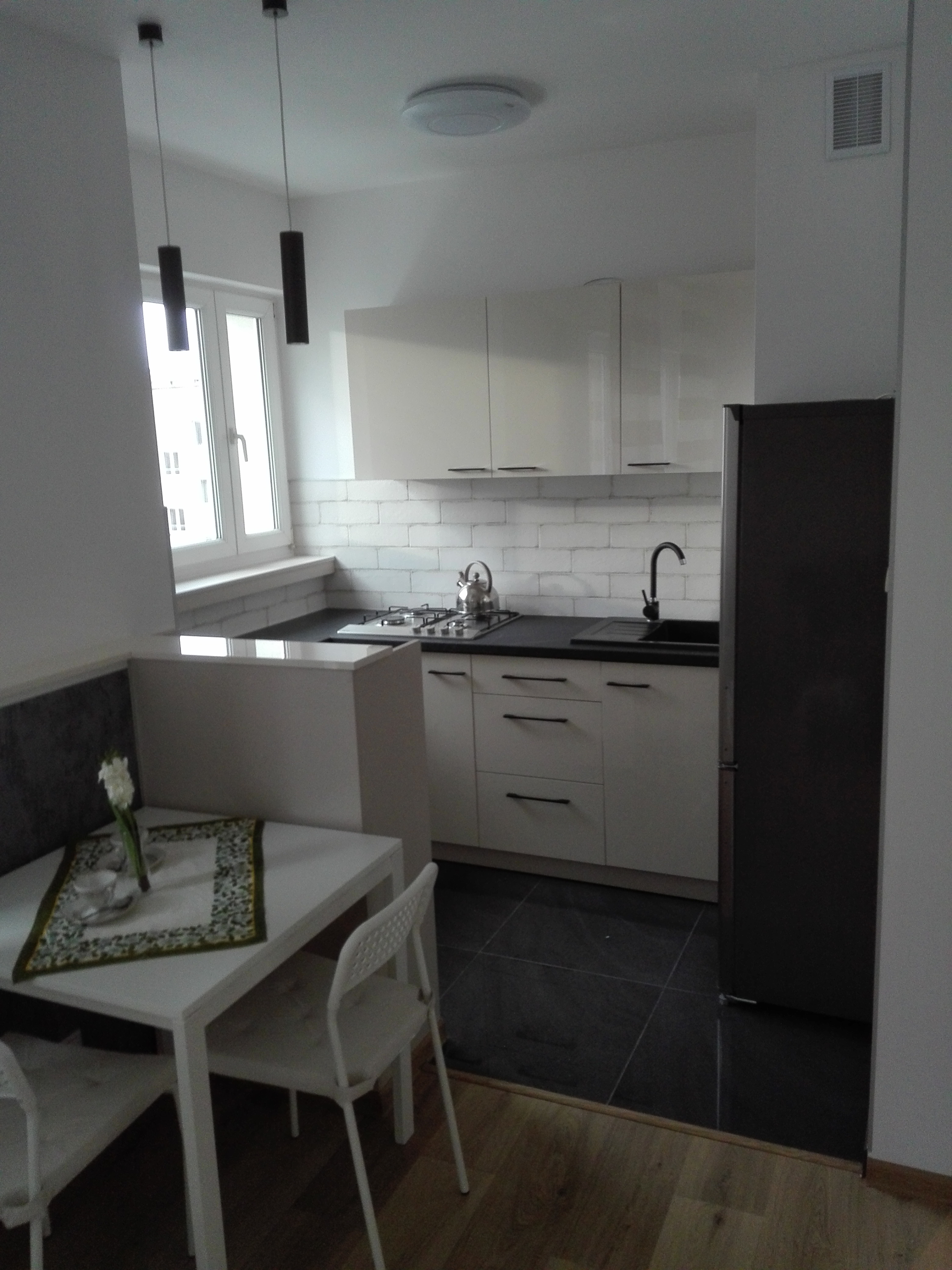 Jasna kuchnia z białymi szafkami, czarnym blatem i płytkami imitującymi cegłę, widok na stół z dekoracyjnym obrusem i kwiatami, minimalistyczne lampy wiszące.