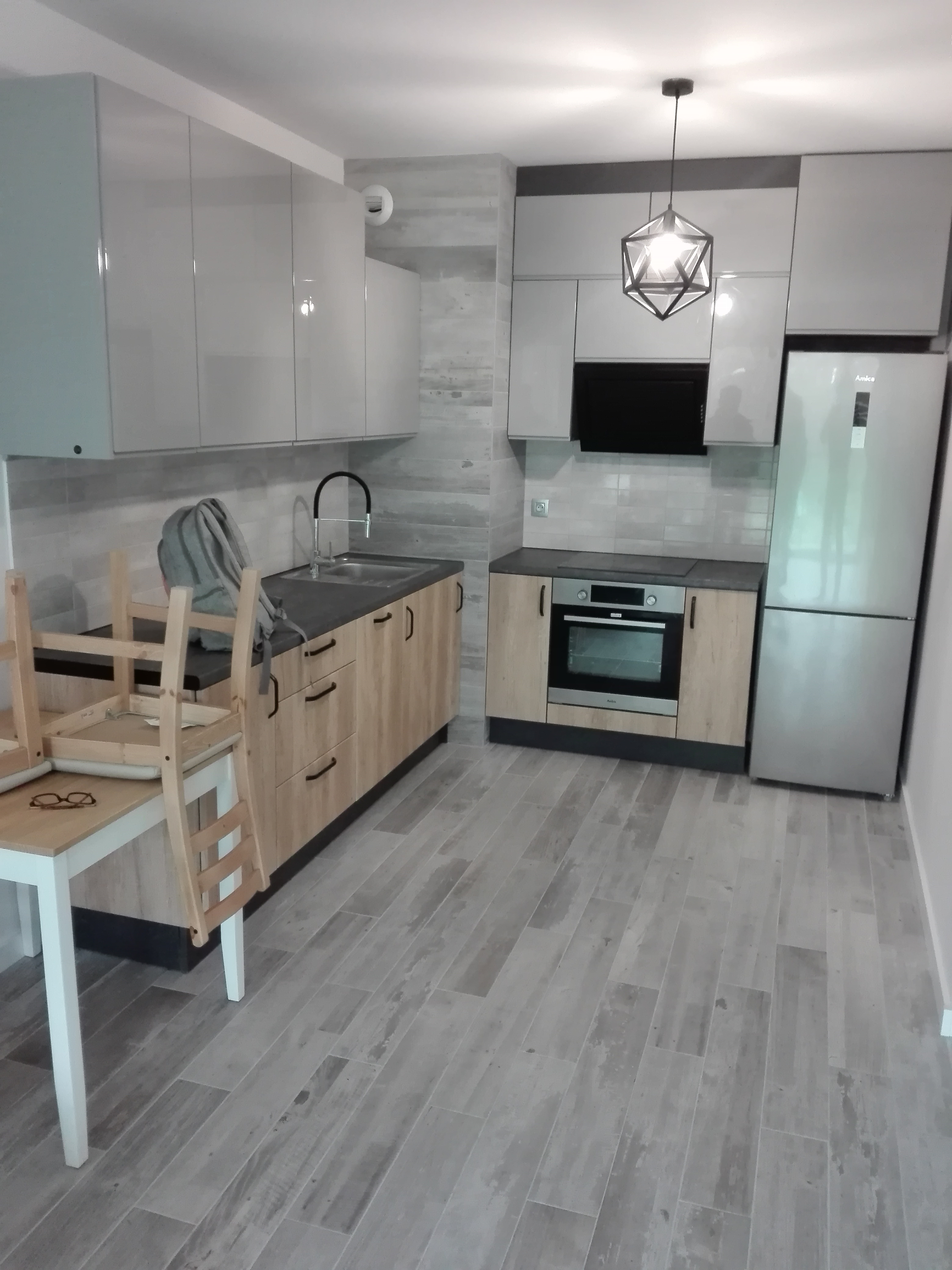 Jasna kuchnia z drewnianymi frontami szafek dolnych i szarymi górnymi, z blatem w kolorze antracytowym, oświetlona geometryczną lampą wiszącą, z krzesłem i okularami na stole w rogu.