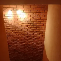 Ściana z cegły dekoracyjnej w korytarzu, oświetlona dwoma żarówkami, z widocznymi fragmentami pomalowanych ścian bocznych i podłogi z płytek.