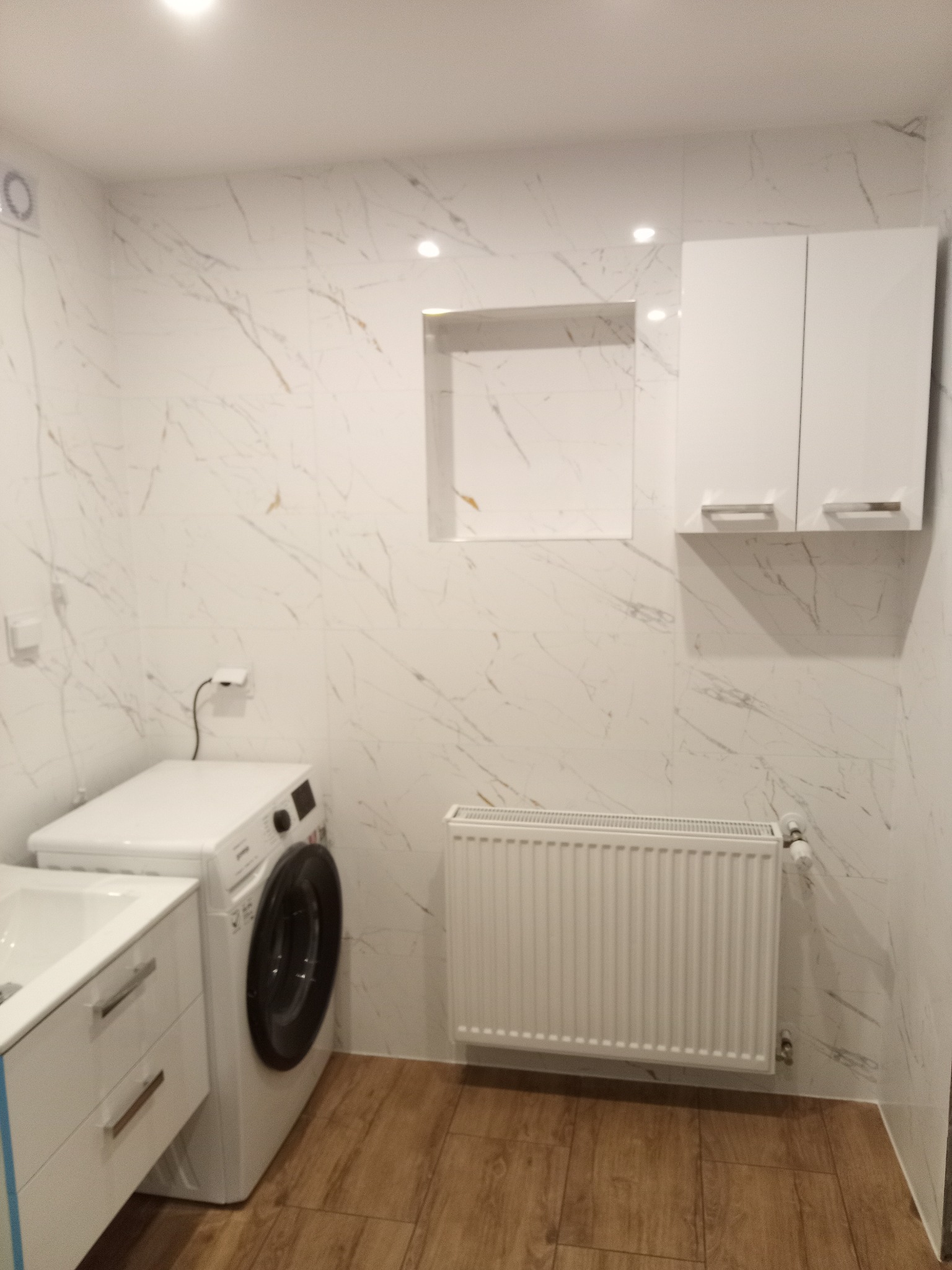 Wykończona łazienka z marmurowymi płytkami, białą szafką, pralką i grzejnikiem. Uwagę zwraca wnęka w ścianie. Podłoga wyłożona panelami imitującymi drewno.