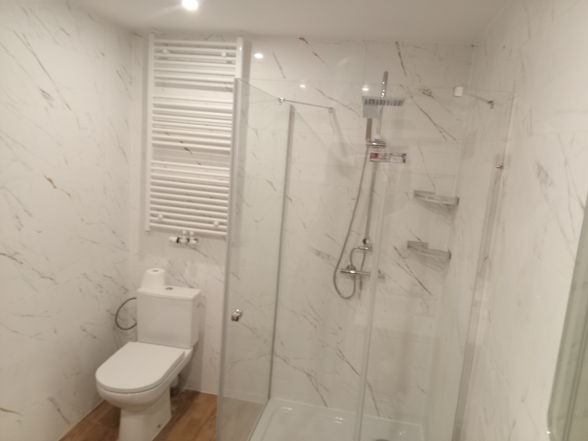 Wykończona łazienka z kabiną prysznicową, toaletą i grzejnikiem drabinkowym. Białe marmurowe płytki na ścianach, drewnopodobna podłoga. Nowoczesny design.