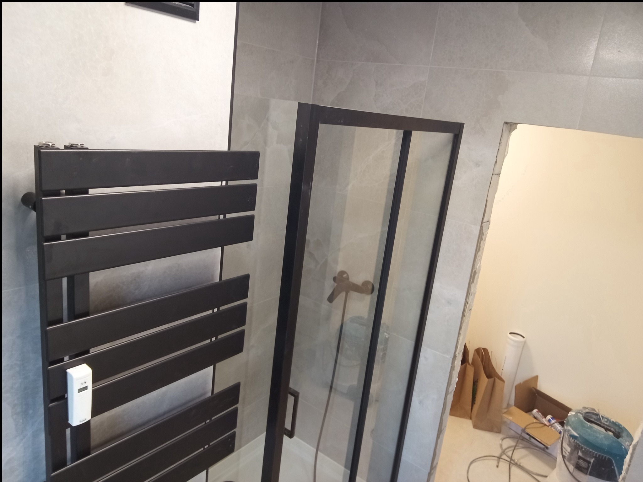 Nowoczesna łazienka w trakcie remontu: czarny grzejnik drabinkowy, kabina prysznicowa z czarnymi ramami i szare płytki. Widoczne narzędzia i materiały budowlane.