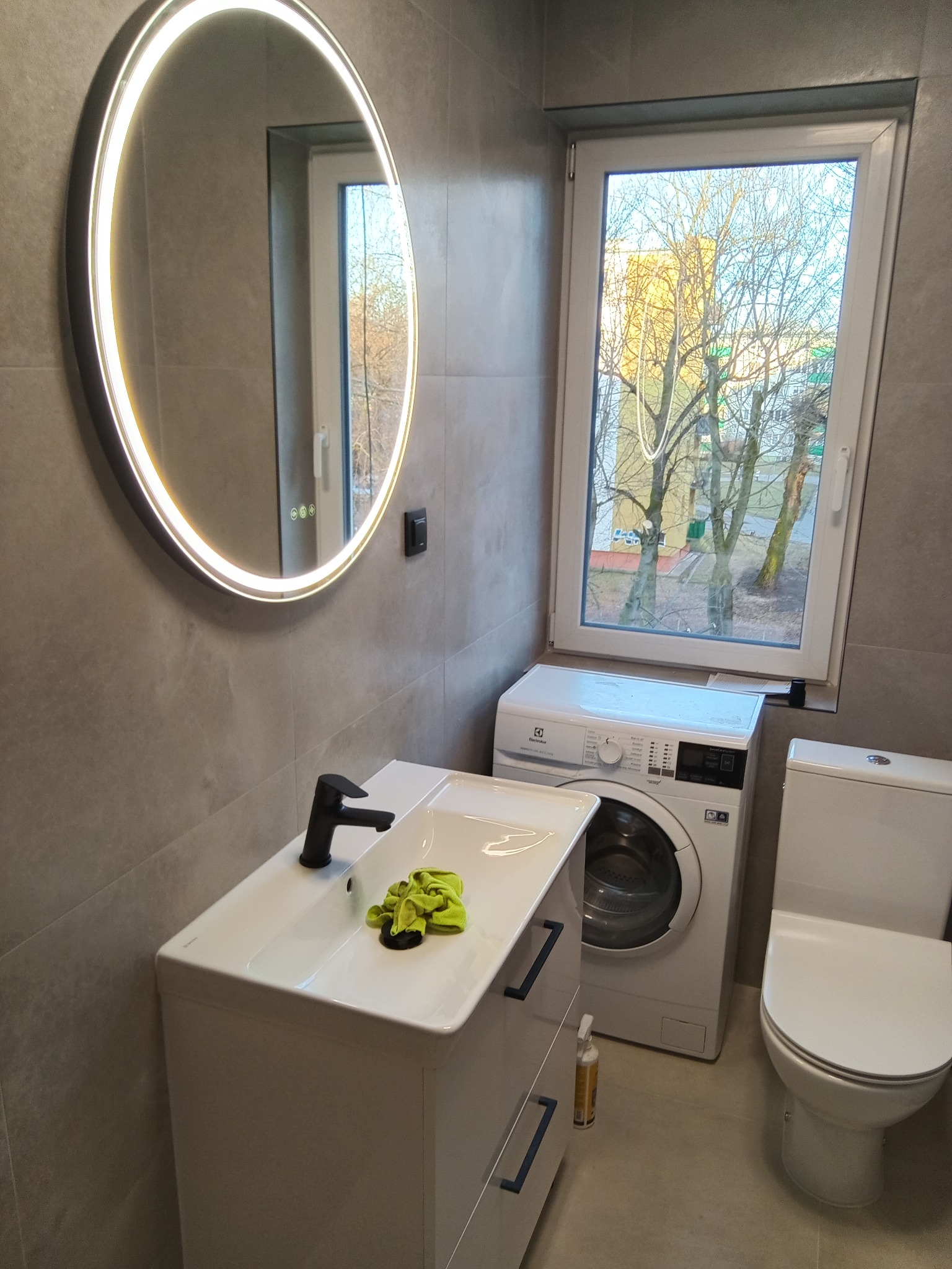 Nowoczesna łazienka w szarościach z okrągłym lustrem LED, białą umywalką z szafką, pralką i toaletą. Widok z okna na drzewa. Minimalistyczny design.