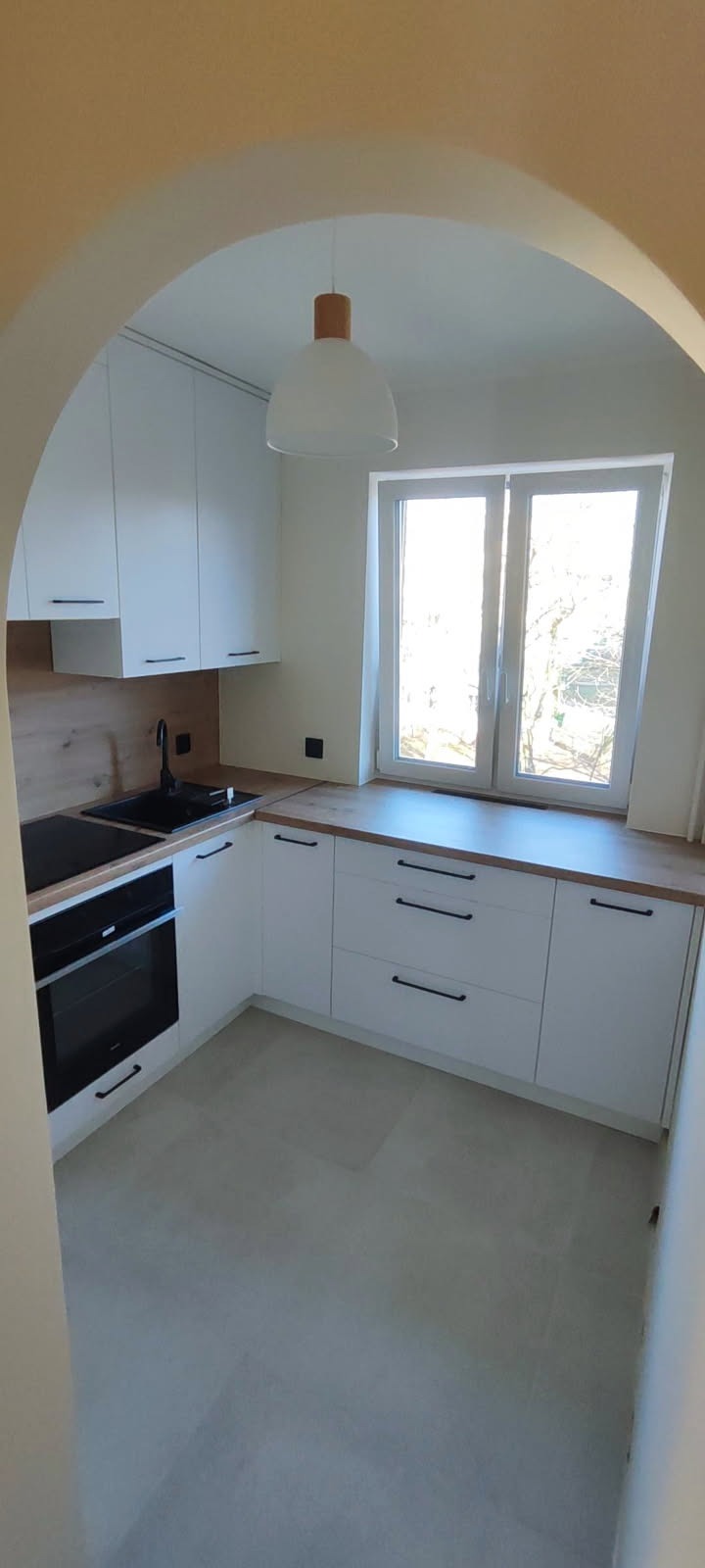 Jasna kuchnia z białymi szafkami i drewnianym blatem, widziana z perspektywy łuku. Minimalistyczny design, czarne uchwyty i armatura. Okno z widokiem na drzewa.