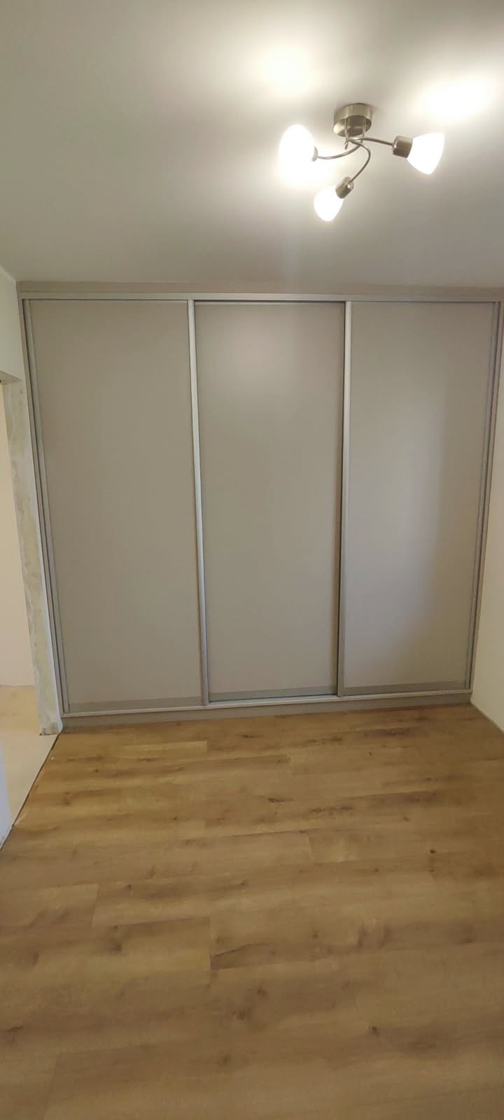 Beżowa szafa przesuwna z trzema drzwiami i drewnopodobną podłogą w jasnym pokoju. Widoczna lampa sufitowa. Minimalistyczny design.