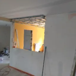 Wnętrze w trakcie remontu: widoczne profile sufitowe, fragment pomalowanej na ż&oacute;łto ściany oraz ściany przygotowane do gładzi, z widocznymi niedoskonałościami i resztkami starej farby.