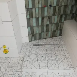 Wykończona łazienka z białymi płytkami z geometrycznym wzorem na podłodze i ścianach, oraz pionowymi płytkami w odcieniach zieleni i szarości w strefie prysznica. Widoczne elementy instalacji...