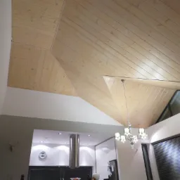 Drewniany sufit pod skosem z widocznymi deskami, oświetlenie halogenowe i żyrandol w nowoczesnym wnętrzu z oknem trapezowym.