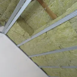 Izolacja poddasza wełną mineralną zamontowana między metalowymi profilami, widoczne drewniane elementy konstrukcyjne i białe sznurki mocujące.