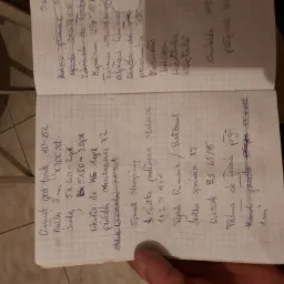 Otwarty notatnik z listą materiałów budowlanych, w tym 'grunt pod tynk', 'siatka podłogowa stalowa', 'wieszak ES', zapisanych niebieskim długopisem na papierze w kratkę.