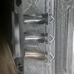 Metalowa forma z trzema półcylindrycznymi wgłębieniami o różnej fakturze powierzchni, przymocowana do metalowego bloku z widocznymi śladami obróbki.