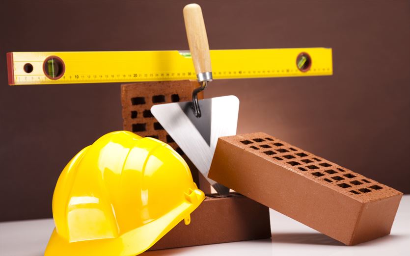 Martwa natura z cegłami, kielnią, poziomicą i żółtym kaskiem budowlanym na białym blacie i brązowym tle.