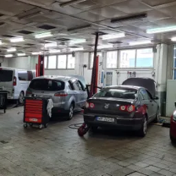 Wnętrze warsztatu samochodowego z kilkoma autami: czerwony Renault Clio z otwartą maską na pierwszym planie, obok ciemny Volkswagen Passat, dalej srebrny Ford S-Max i biały Opel Vivaro, a w tle...