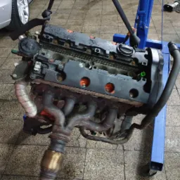 Zdemontowany silnik samochodowy zawieszony na niebieskim stojaku warsztatowym, widoczne kolektory wydechowe, głowica i część bloku silnika, tło stanowi podłoga warsztatu i fragment nadkola...