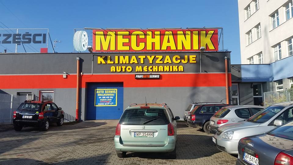 Budynek warsztatu samochodowego z dużym szyldem 'Mechanik Klimatyzacje Auto Mechanika', zaparkowane samochody przed budynkiem, błękitne niebo.