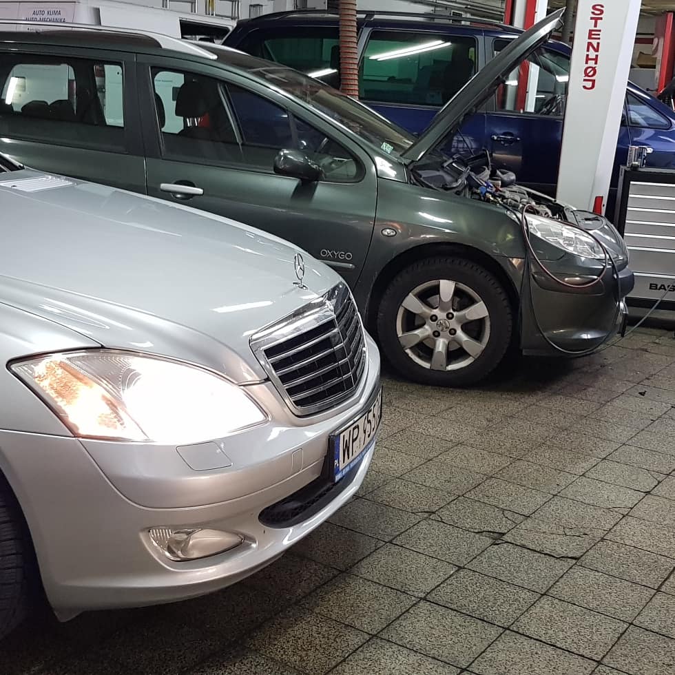 Dwa samochody w warsztacie: srebrny Mercedes z przodu i ciemnozielony Peugeot z otwartą maską, podłączony do urządzenia diagnostycznego.