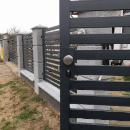 Nowoczesna brama wjazdowa z metalowymi przęsłami i okrągłą gałką, dopasowana do ogrodzenia z betonowymi słupkami i metalowymi panelami, widok z zewnątrz na tle pochmurnego nieba.