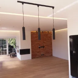 Jasne wnętrze po remoncie z parkietem, ceglaną ścianą z kominkiem i nowoczesnym oświetleniem sufitowym z trzema lampami cylindrycznymi