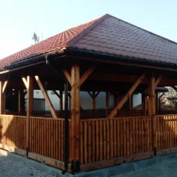 Drewniana altana ogrodowa z brązowym dachem z blachodachówki, drewnianą balustradą i widocznymi rynnami, na szarej nawierzchni.