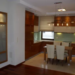 Nowoczesna kuchnia z drewnianymi szafkami, białymi frontami i stołem jadalnym z krzesłami w kolorze ecru, oświetlona wiszącymi lampami i punktowymi światłami w suficie.