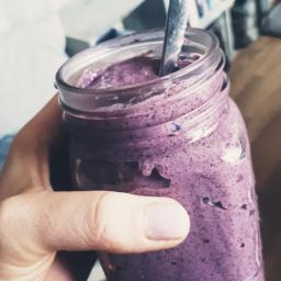 Marta Bąkowska - Fioletowy koktajl jagodowy w słoiku trzymanym w dłoni, z łyżeczką w środku, na tle regału z książkami.