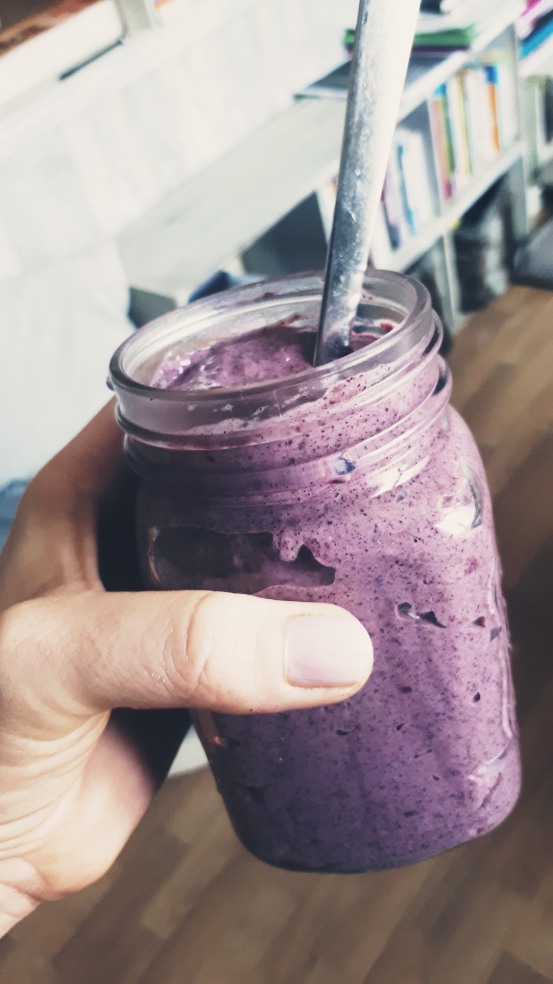 Fioletowy koktajl jagodowy w słoiku trzymanym w dłoni, z łyżeczką w środku, na tle regału z książkami.